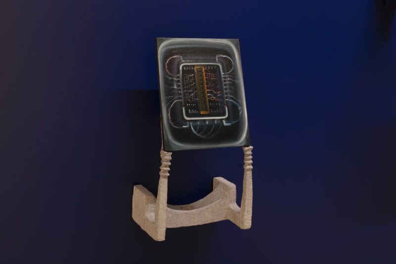 A painting of an electronic circuit board is displayed upright on a two-pronged beige stand against a dark blue background.