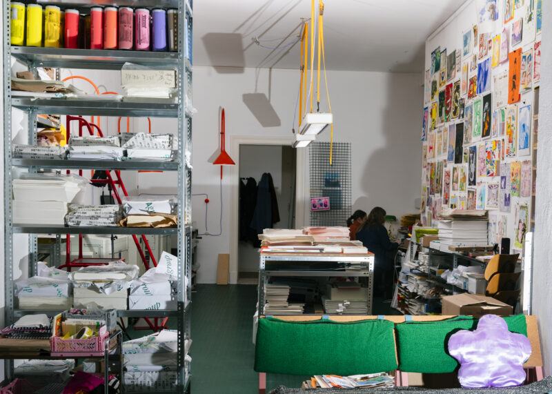 Colorama project space with shelving on the left containing reams of paper, a table sits in the middle of the room, and a shelf to the right sits full of supplies under a wall covered in colorful illustrations.