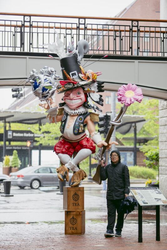 A person stands next to a large, colorful whimsical statue, holding a flower and rifle, set outdoors on wooden tea crates.
