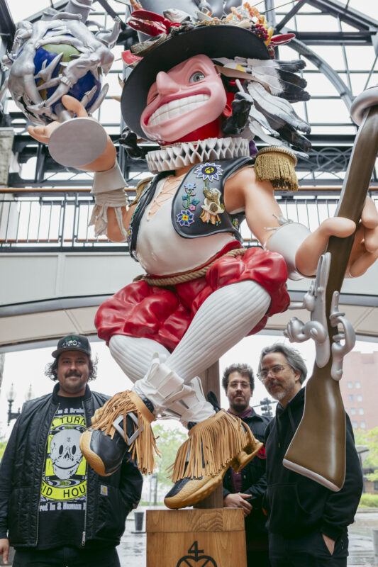 Three men stand in front of a large, colorful statue of a smiling figure, holding a staff and globe.