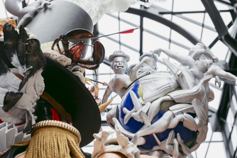 A close-up of a surreal sculpture featuring human figures coming out of a globe, a lobster, inside a building with a glass ceiling.
