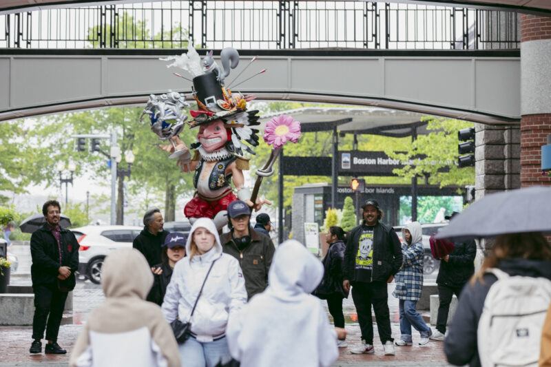 A crowd of people walk past a a large colorful statue of an exaggerated figure wearing a top hat with various colorful objects on it, and holding a silver globe with figures coming off of it.