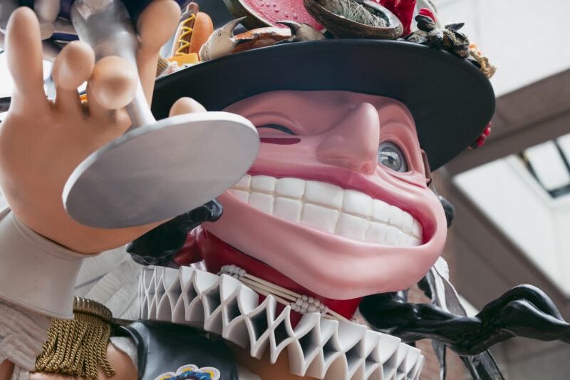 Close-up of a large, colorful statue with exaggerated facial features, wide grin, winking eye, and an elaborate hat adorned with various objects.