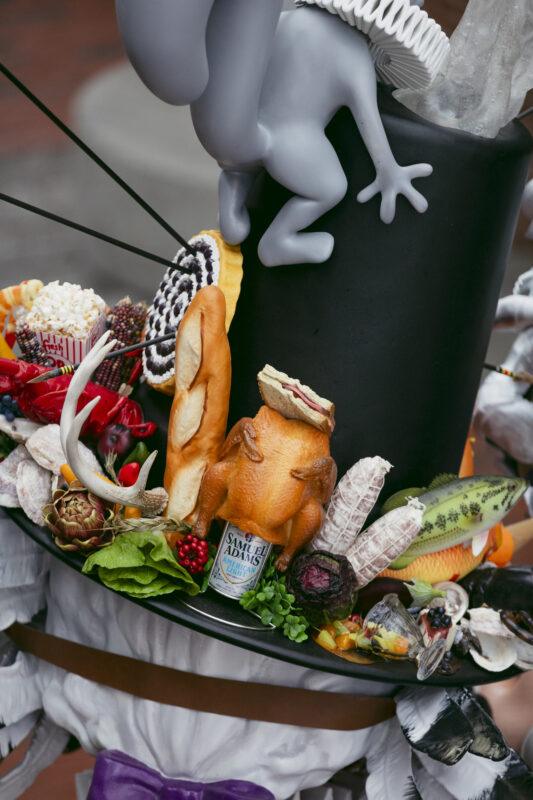 A black hat decorated with a faux gray animal, bread, vegetables, a small canned food, feathers, and other eclectic items.