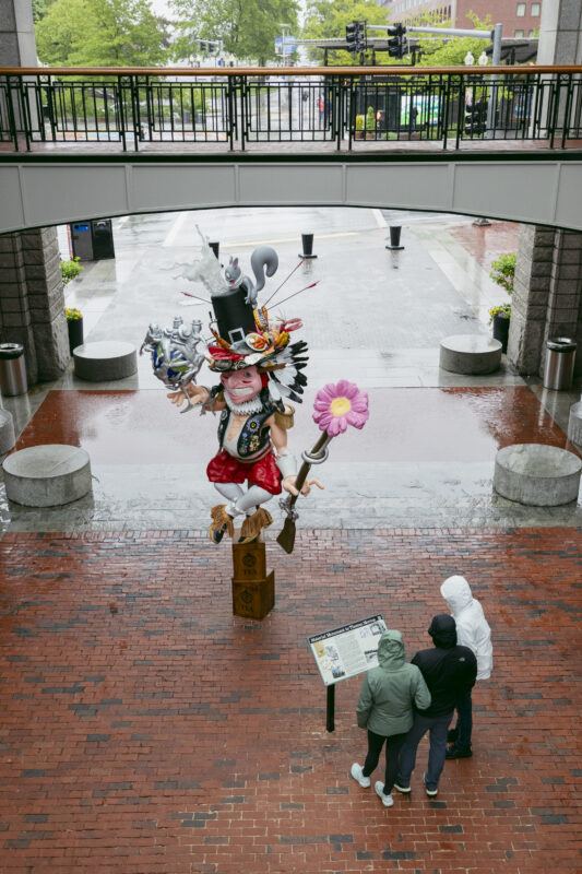Three people in hooded jackets read a sign near a colorful, whimsical statue on a rainy brick plaza under an overpass.