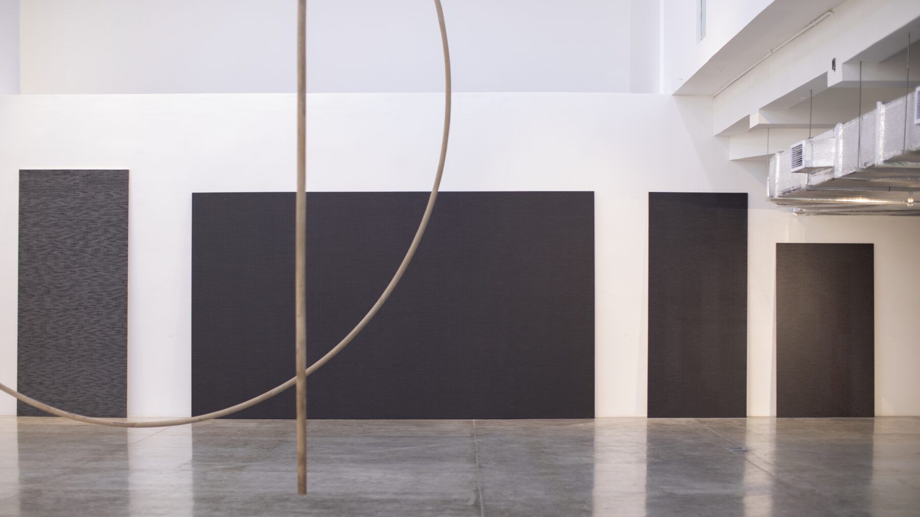 Four large, rectangular black panels are mounted on a white wall in a gallery space with polished concrete floors and a curved sculpture in the foreground.