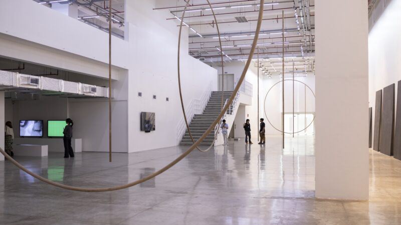 A spacious modern art gallery with large circular sculptures, wall-mounted artworks, and a few people observing the exhibits.
