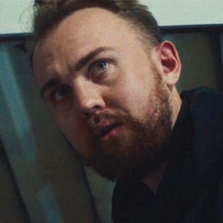 Trey Abdella with a beard and short hair looks up with a serious expression, standing indoors near a corrugated metal ceiling.