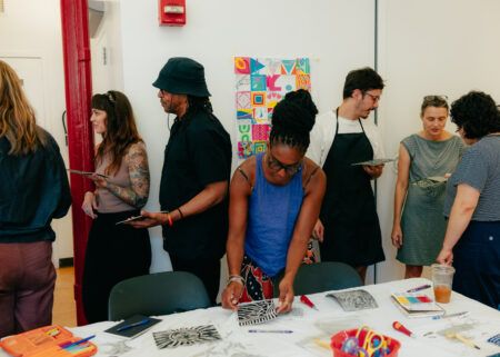 People participate in an art workshop, creating and examining artwork on tables and walls in a brightly lit room.