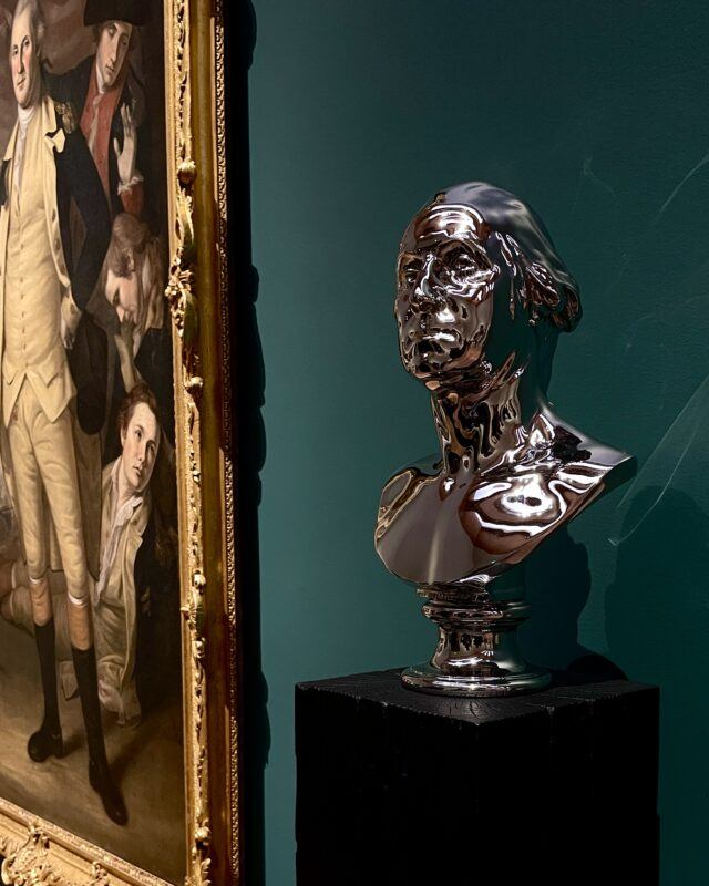 A polished metallic bust on a pedestal is displayed beside a framed historical painting against a dark green wall.