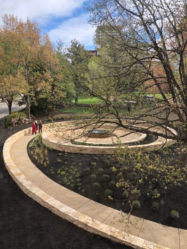 A circular stone walkway surrounds a small garden and central fountain, with three people walking along the path. Tall trees with budding leaves frame the scene.