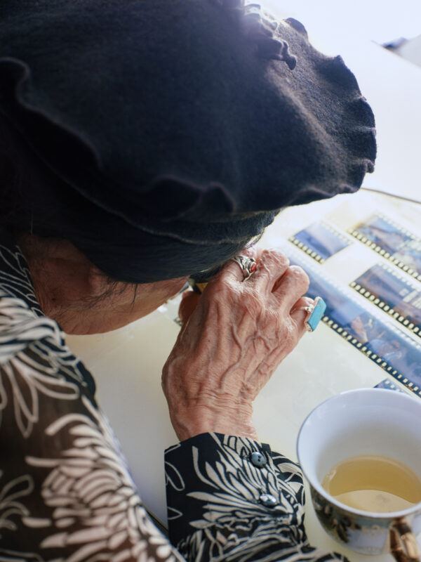 Alanis Obomsawin examines film negatives with a magnifying glass at a table, next to a cup of tea.