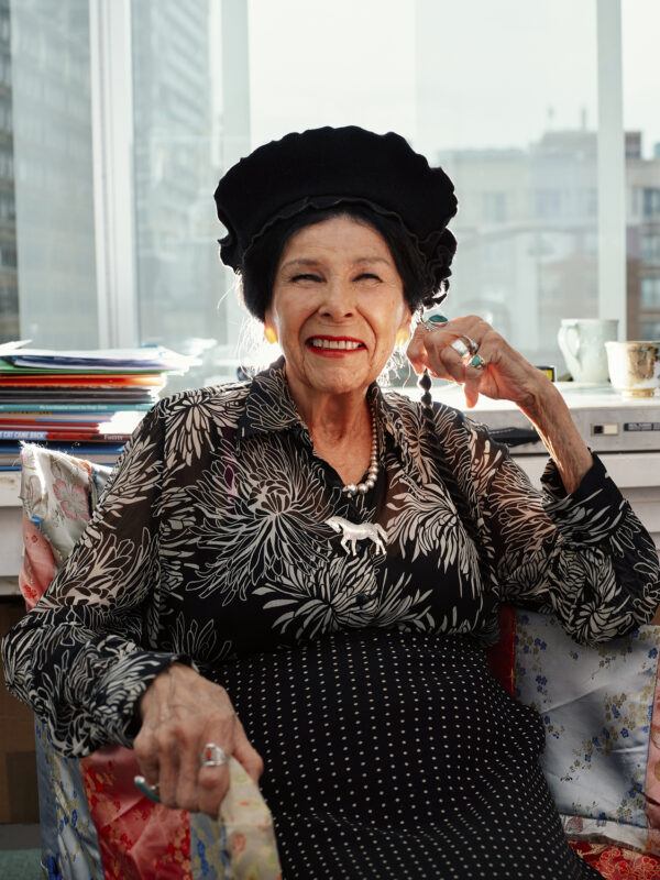 Alanis Obomsawin sits in a colorful chair by a window, smiling, wearing a black turban, patterned blouse, polka dot skirt, and several rings. Stacks of papers and cups are visible in the background.