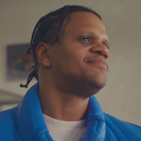 American Artist with braided hair and wearing a blue jacket looks off to the side and smiles slightly indoors.