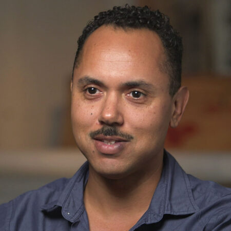 Edgar Arceneaux with short curly hair and a mustache, wearing a blue collared shirt, sits indoors with a neutral background.
