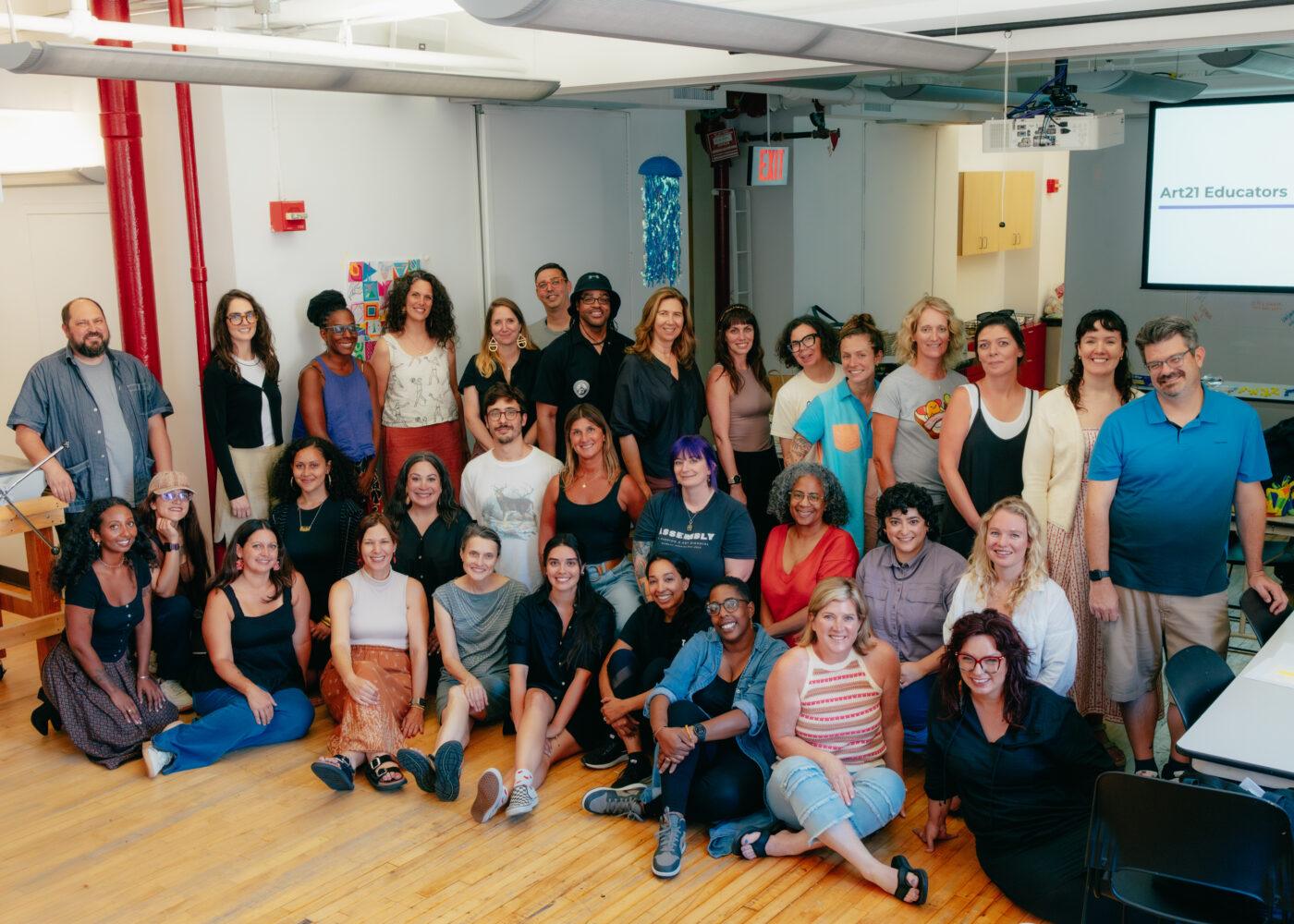 A diverse group of adults pose and smile for a group photo in a well-lit classroom or workshop setting with wooden floors.