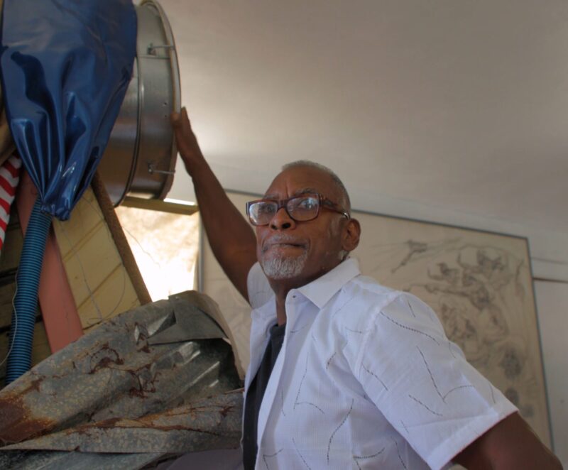Daniel Lind-Ramos in glasses and a short-sleeve button-up shirt stands indoors, reaching up to a large metallic drum coming out of a tall sculpture.