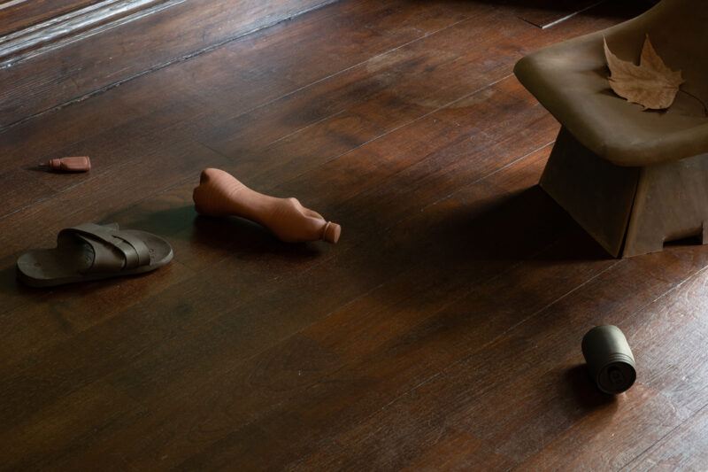 Four clay sculptures resembling discarded items like a plastic bottle or a single sandal are arranged on a dark wooden floor.