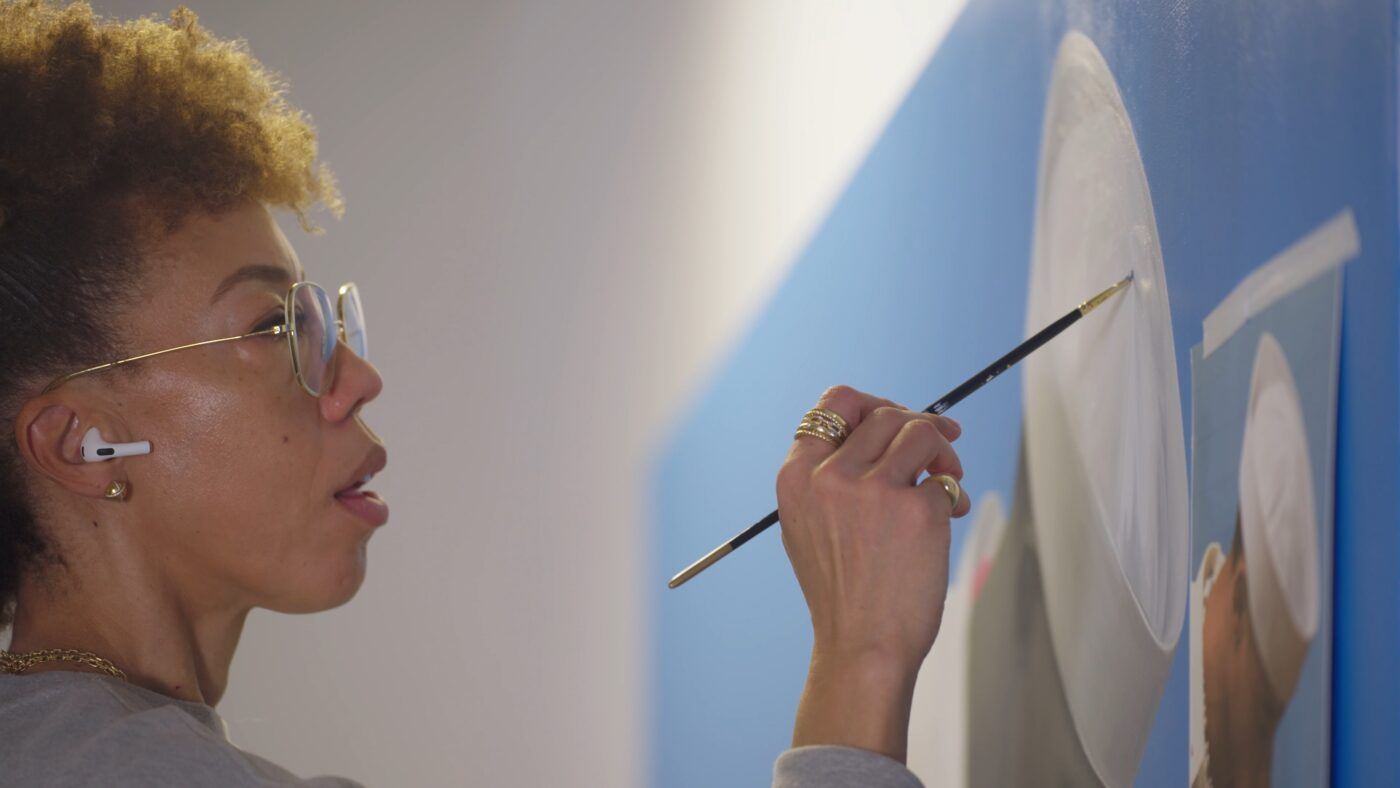 Amy Sherald wearing glasses and earbuds paints on a canvas with a thin brush, with a focused expression.