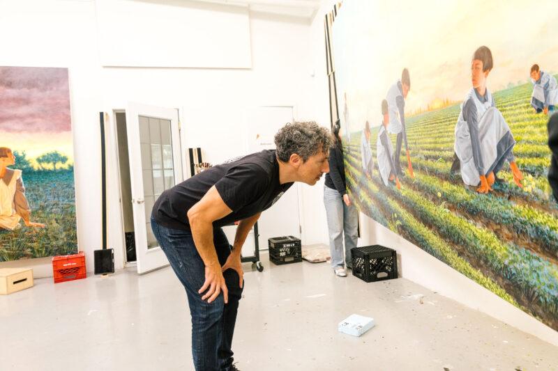 Kent Monkman leans forward to examine a large, detailed painting of people working in a field, displayed in an art studio.