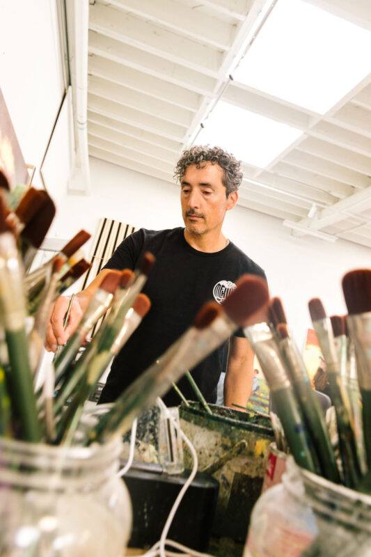Kent Monkman stands in his studio, reaching toward a surface, with jars of paintbrushes visible in the foreground.