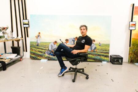 Kent Monkman sits on a chair in an art studio holding a paintbrush, with a large painting of people in a field displayed behind him and art supplies nearby.