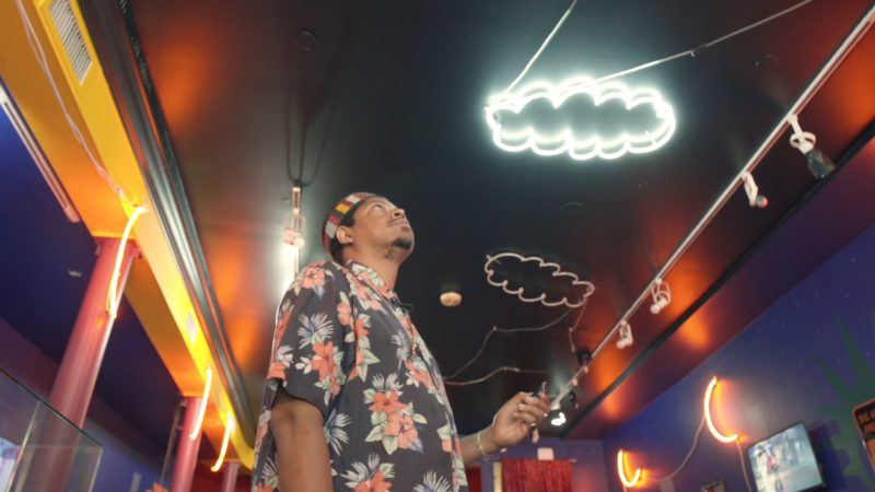 Azikiwe Mohammed wearing a floral patterned shirt, standing in a purple room with yellow beams, with red support poles, looking up at a black ceiling with neon clouds on the ceiling.