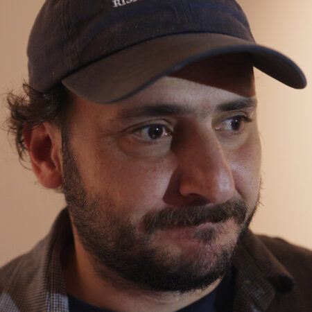Neïl Beloufa with a beard and mustache wearing a dark baseball cap looks to the side with a neutral expression.
