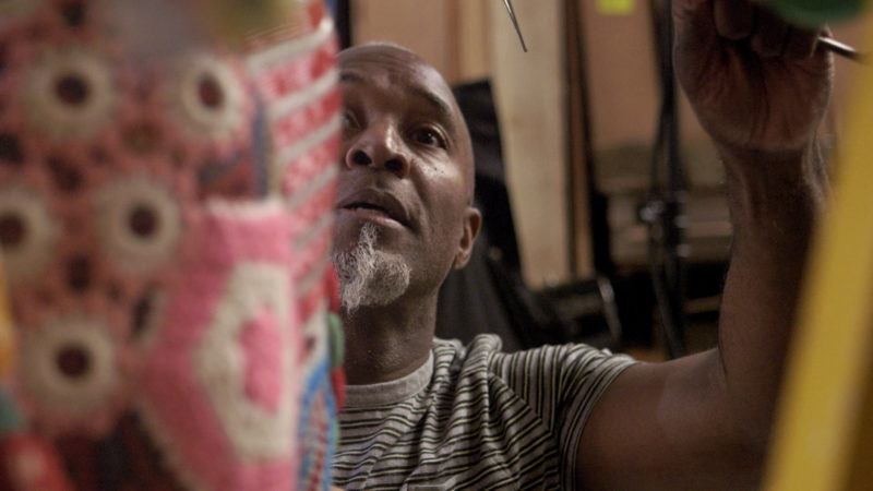 Nick Cave working on a fabric piece, the piece is out of focus in the foreground of the photo, Cave's face peaks through while is arm is raised to work on the piece.