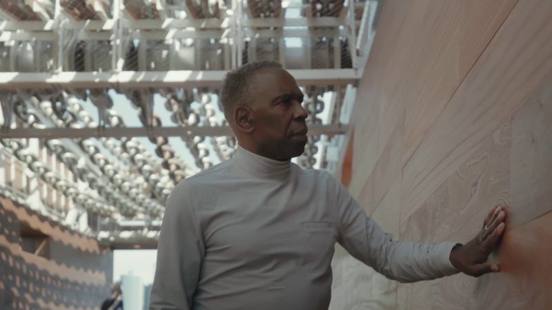 Charles Gaines standing inside his sculpture "Moving Chains." He places his hand on the wall next to him, and his looking up at the oversized chains above.