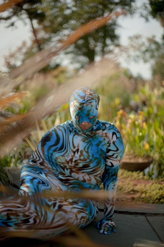 Person in a blue, brown, and white patterned bodysuit sitting outdoors near plants and stone pathway.
