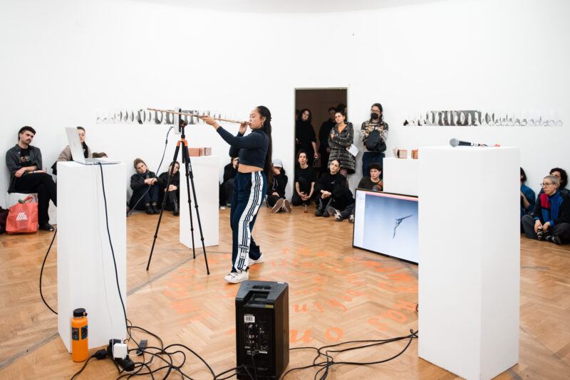 Caroline Garcia stands in a gallery performing with a blowgun as people sit and watch; art installations and a monitor surround her.