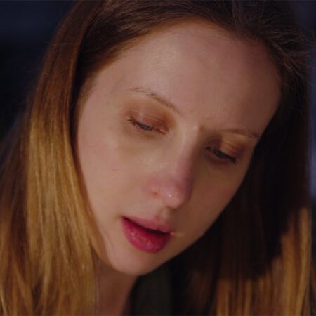 Sara Cwynar with long light hair looks downward with a serious expression in soft indoor lighting.