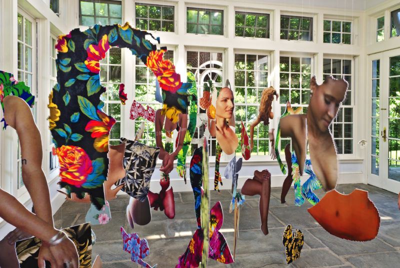 Hanging cut outs of abstract human figures with floral patterns suspended in a sunlit room with large windows.