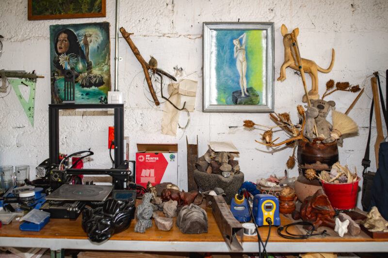 A cluttered worktable with artworks, tools, a 3D printer, and sculptures.