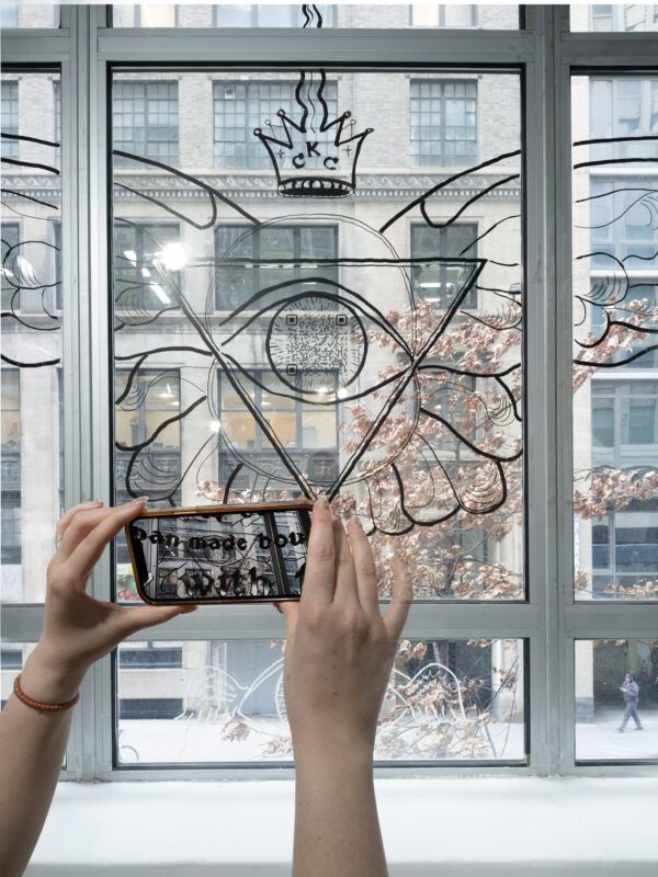 Two hands hold a phone to take a photo of a large eye illustration with wings and a crown drawn on a window, with a city building visible outside.