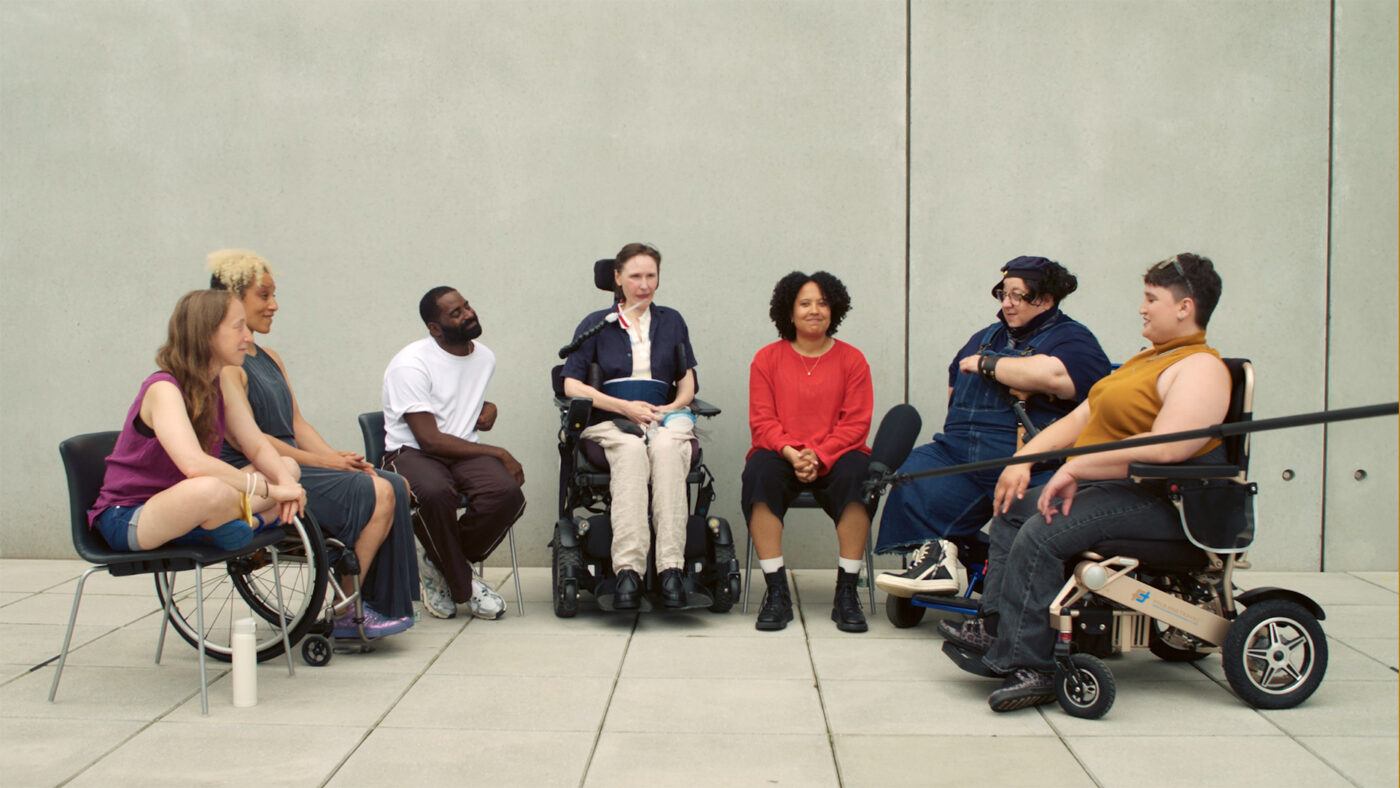 A diverse group of seven people, some using wheelchairs, sit in a semicircle outside against a plain concrete wall, engaged in conversation.
