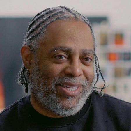Arthur Jafa with braided hair, a beard, and an eyebrow piercing smiles while wearing a black shirt.