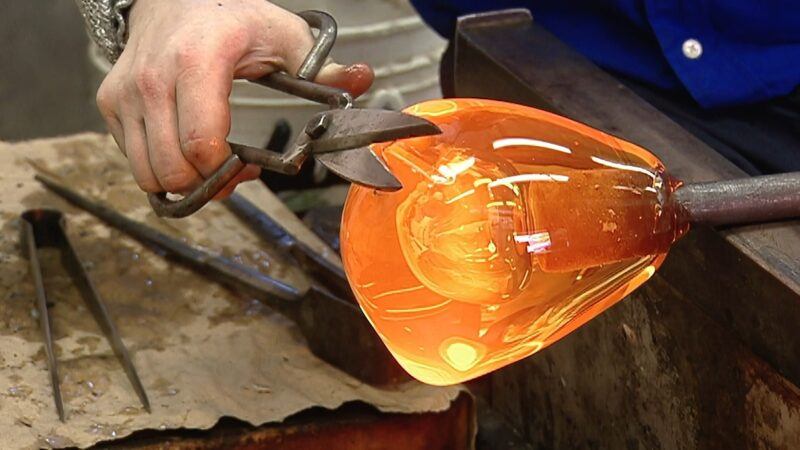A person shapes hot molten glass with metal tools during the glassblowing process.
