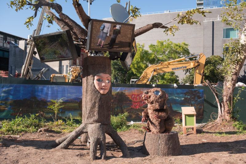 Tree sculpture with a human face and television screens. coming out of the top of it, next to a beaver sculpture and construction equipment in an outdoor urban setting.