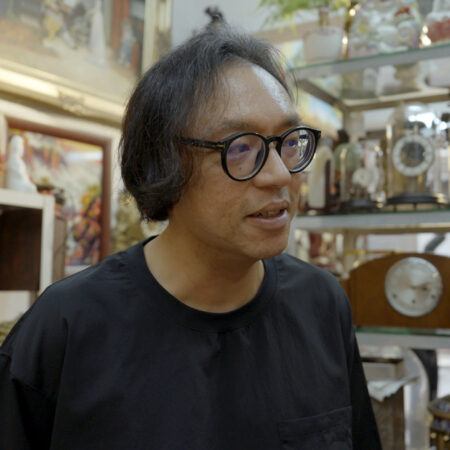 Ho Tzu Nyen with medium-length dark hair and glasses stands indoors, surrounded by shelves displaying various vintage clocks and decorative items.