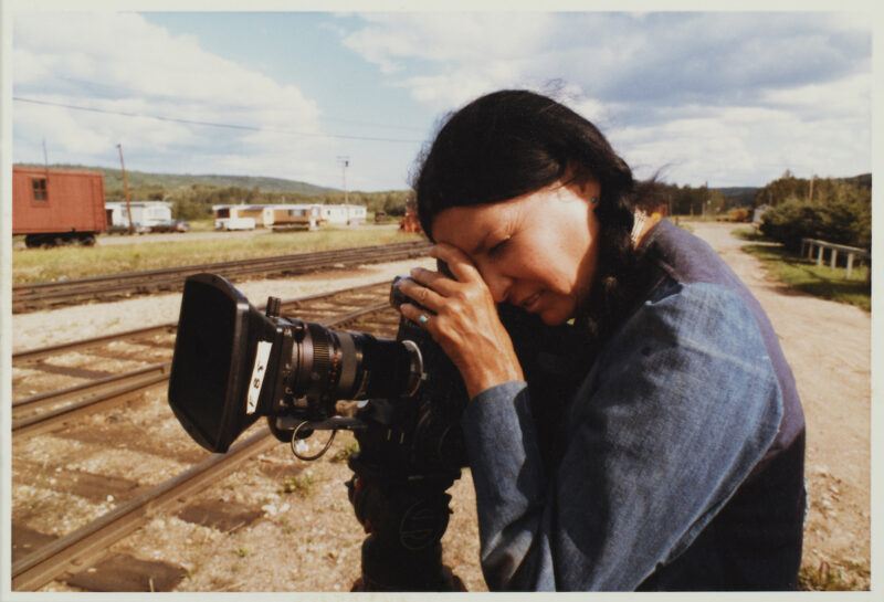 A young Alanis Obomsawin with long dark braids operates a film camera beside railroad tracks, with train cars and trees visible in the background under a partly cloudy sky.
