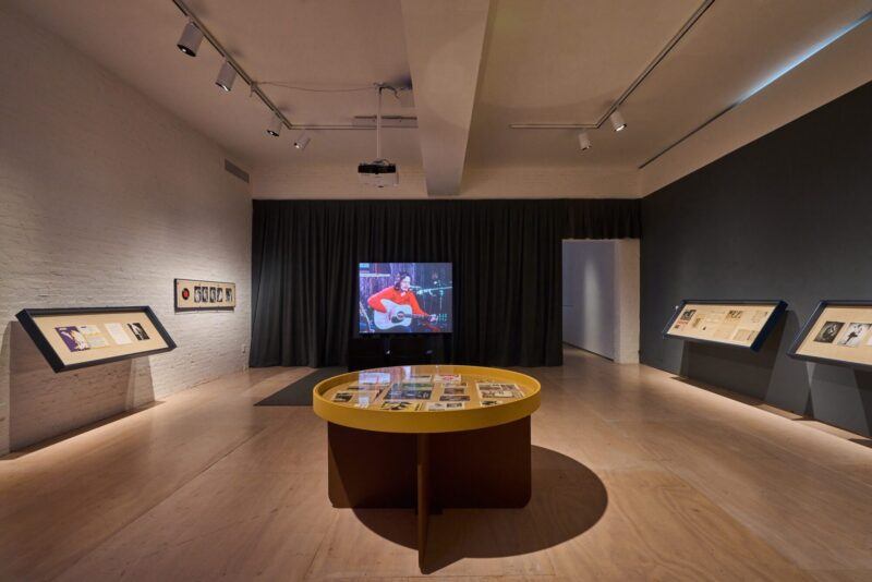 A gallery room displays framed documents and photos on slanted stands, with a round table in the center and a screen showing a person playing guitar in the background.