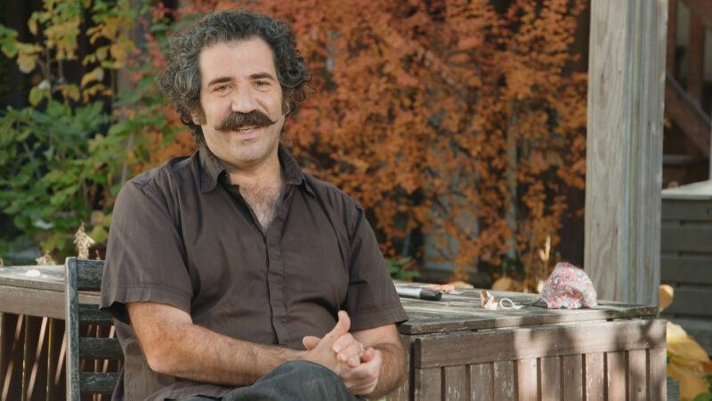 Michael Rakowitz with curly hair and a mustache sits outdoors on a wooden bench, with autumn foliage in the background.