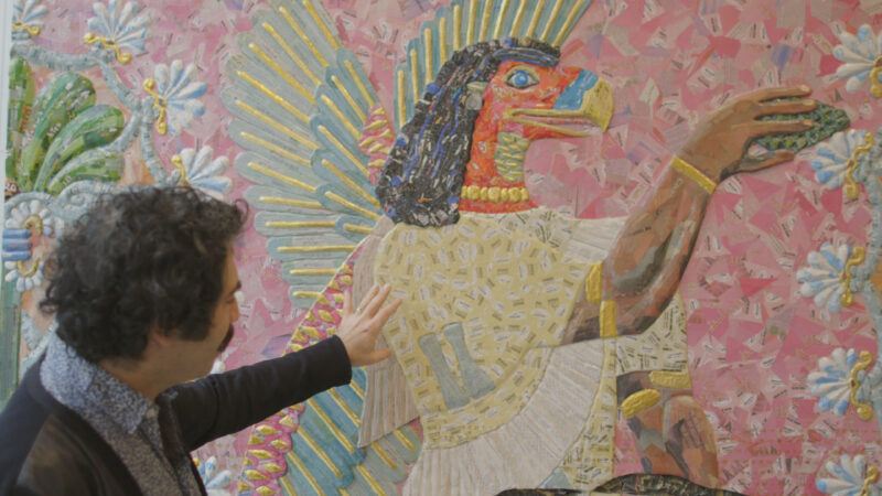 Michael Rakowitz gestures toward a colorful mosaic depicting a winged, bird-headed figure against a patterned background.
