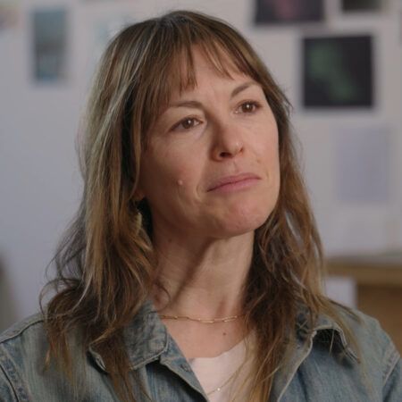 Lucy Raven with shoulder-length hair and bangs wears a denim jacket and white shirt, sitting indoors with a blurred wall of photos and a table in the background.