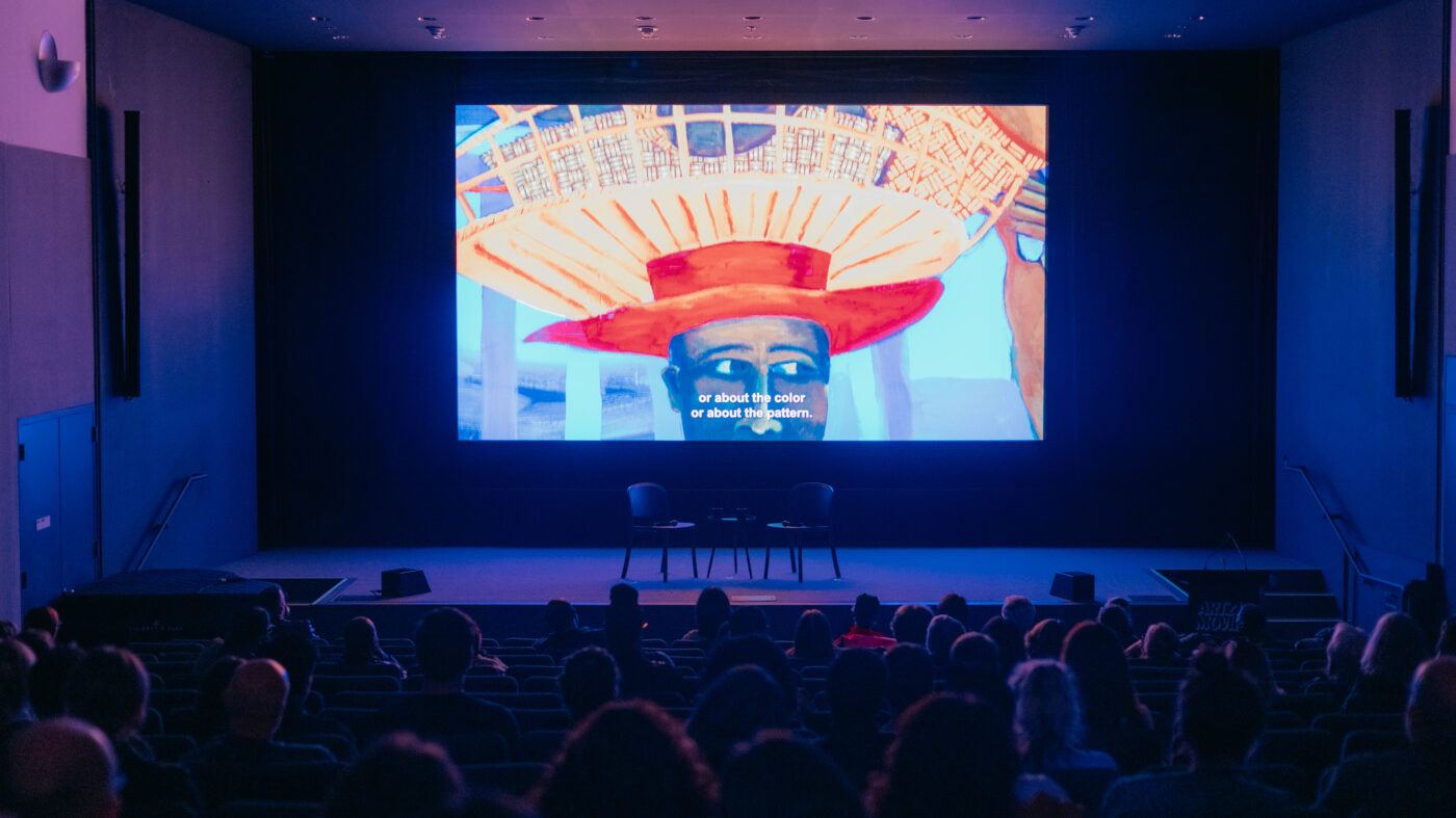 Audience seated in a dark theater watching a film with vibrant artwork on screen and English subtitles visible. Two empty chairs are on the stage in front of the screen.