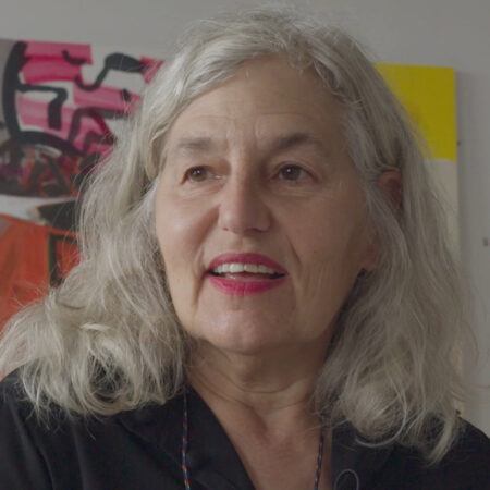 Amy Sillman with shoulder-length gray hair wearing a black blouse stands in front of colorful abstract paintings.