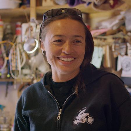 Rose B. Simpson smiling out past the camera with sunglasses atop her head, her studio is out of focus in the background.