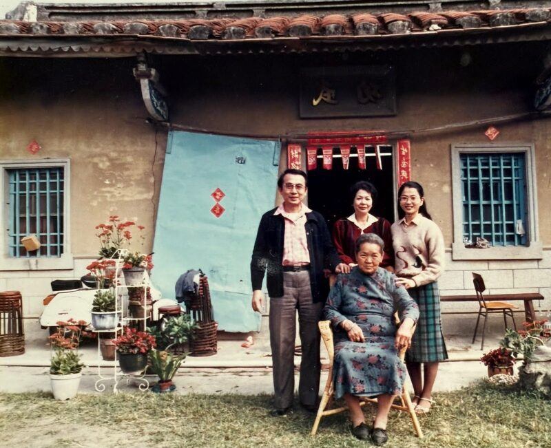 Four people pose in front of a house with blue window bars and red decorations; three stand behind an elderly woman seated in a chair.